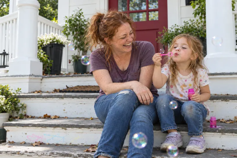 A mom laughing on her front porch steps as her daughter blows soap bubbles