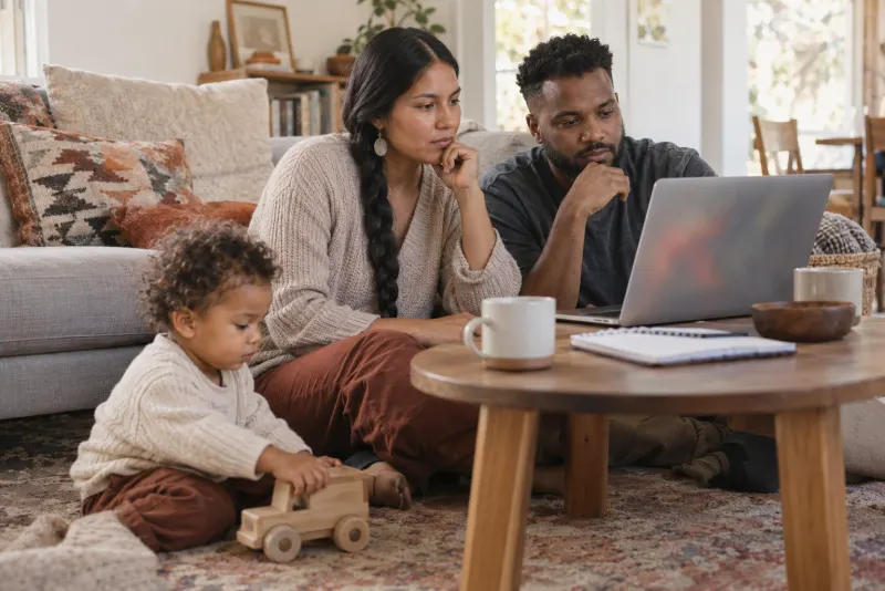 Parents sitting on the living room rug comparing options on a laptop while their toddler plays with a wooden truck